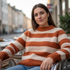 May include: A woman wearing a rust and cream striped sweater with a mock neck. The sweater has long sleeves and is made of a knitted material. The woman is sitting outside on a sunny day.