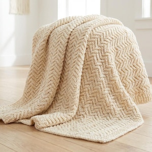 Può includere: Una coperta beige lavorata a maglia con un motivo a spina di pesce è drappeggiata su un pouf arrotondato. La trama e il design della coperta sono visibili, con il tessuto che scende lungo i lati. La stanza ha una palette di colori neutri.