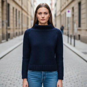May include: A navy blue sweater with a high neckline and long sleeves. The sweater is paired with blue jeans. The person is standing on a cobblestone street with buildings on either side.