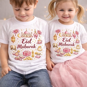 May include: Two children wearing white t-shirts with a colourful "Eid Mubarak" design. The design includes a mosque, crescent moon, lanterns, and gifts. The children are wearing blue jeans and a pink skirt.