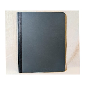 May include: A rectangular notebook with a dark gray textured cover and a contrasting black vertical spine strip. The front cover features a subtle grid-like pattern across its surface, while the spine has a smooth, dark, and slightly reflective finish.