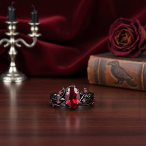 Peut inclure: Bague noire ornée d'une grande pierre rouge ovale et de petites pierres rouges. La bague présente un design sombre et orné avec un motif floral ou en forme de vigne. La bague est posée sur un fond sombre.