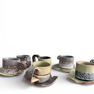 May include: A collection of handmade ceramic espresso cups and saucers. The cups feature a variety of colors, including gray, green, and brown, with geometric patterns in black and white. The saucers are leaf-shaped and complement the cups.