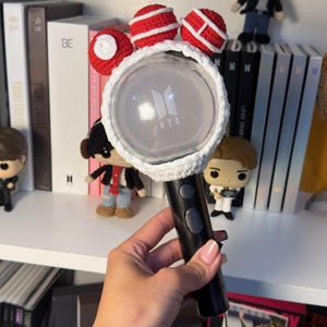 Op de afbeelding: Een zwarte BTS-lichtstick met een wit en rode gehaakte hoes. De hoes heeft drie rode bollen met witte strepen. De lichtstick toont het BTS-logo en is omgeven door andere BTS-merchandise.
