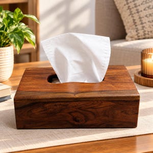 May include: A rectangular tissue box cover crafted from dark brown wood with a visible natural grain pattern. The box features a central oval opening on the top surface, through which a single white paper tissue is pulled upward and displayed.