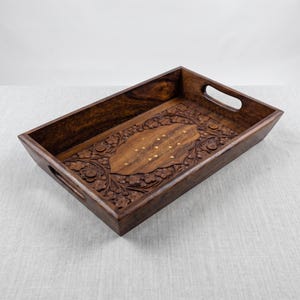 Puede incluir: Una bandeja de servicio rectangular de madera marrón oscuro con intrincadas tallas florales en la base y los lados. El centro de la bandeja muestra un panel liso y alargado incrustado con pequeños detalles florales metálicos claros.