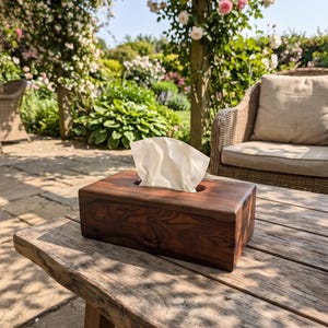 May include: A rectangular tissue box cover crafted from dark brown wood with a visible natural grain pattern. The box features a smooth, polished finish and a central oval opening at the top, from which a single white paper tissue is protruding.