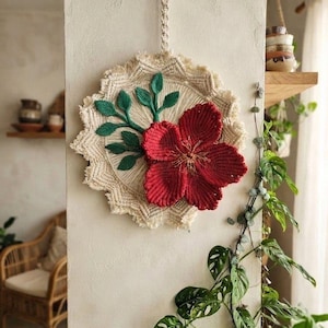 Puede incluir: Un tapiz de pared circular de macramé que presenta una gran flor de color rojo intenso con cinco pétalos prominentes y un centro de fibras finas. La flor está decorada con hojas de macramé verde oscuro sobre una base anudada color crema.