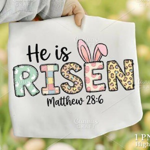 May include: A white t-shirt with the words "He is Risen" in a colorful, decorative font. The letter "R" has pink bunny ears. The text "Matthew 28:6" is below the main text. The shirt is held by a person.