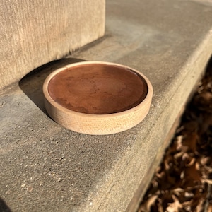 May include: A small, round wooden tray with a dark brown interior. The tray is made of light-colored wood and sits on a concrete surface. The tray's design is simple and minimalist.