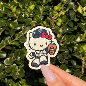 May include: A sticker featuring Hello Kitty dressed as a baseball player. She wears a navy blue cap with a red bow and a baseball glove. The sticker has a white background and the text "MARINERS" on the uniform.