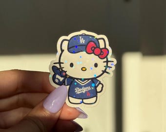 May include: A holographic sticker featuring Hello Kitty in a Los Angeles Dodgers baseball uniform. The sticker shows Hello Kitty wearing a blue cap with the Dodgers logo, a blue jersey, and a red bow. The sticker is held by a hand with purple nail polish.