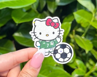 May include: A holographic sticker featuring a white cat character wearing a green soccer jersey with the number 7 and the word MEXICO printed on it, accented by a red bow, positioned next to a black and white soccer ball graphic.