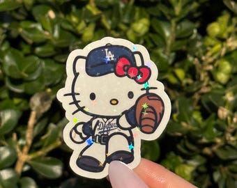 May include: A sticker featuring Hello Kitty in a Los Angeles Dodgers baseball uniform, holding a baseball and glove. The design includes a blue cap with the Dodgers logo, a red bow, and sparkling accents. The sticker is set against a green background.