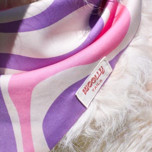 Op de afbeelding: Hondenbandana met een retro golvend patroon in roze, paars en wit. De bandana heeft een label met de tekst "WOOLLY TAILS". De stof is zacht en lijkt zijde of satijn te zijn. De bandana zit om de nek van een hond.