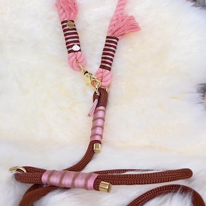 Op de afbeelding: Een meerdelige touwlijn met een roze bovendeel en een bruin onderste deel. De lijn is voorzien van goudkleurige metalen hardware, decoratieve bruin en roze gestreepte draadwikkelingen, kleine kwastjes en een kleine wolk-vormige bedel.