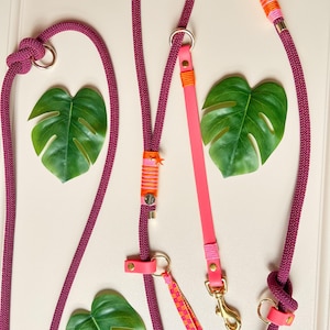 Op de afbeelding: Een donker magenta touwlijn met goudkleurige metalen hardware, een felroze leren riemsectie en decoratieve neonoranje en roze draadwikkelingen. De lijn is voorzien van een kleine sterbedel en een geruit stoffen kwastje als accent.