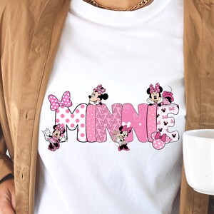 Puede incluir: Camiseta blanca con el nombre "MINNIE" en rosa. Las letras est&aacute;n decoradas con lunares, lazos y personajes de Minnie Mouse. La camiseta la lleva una persona.