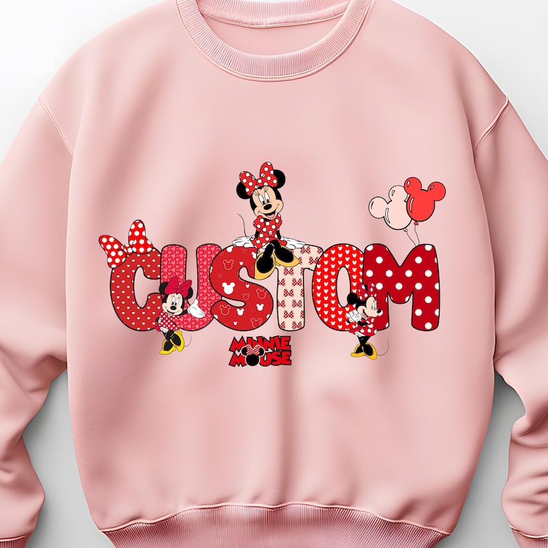 May include: A light pink crewneck sweatshirt featuring a large graphic of the word CUSTOM in red and white polka dot and heart patterns. Minnie Mouse characters are positioned around the letters, accompanied by red and white balloons.
