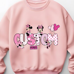 Puede incluir: Sudadera rosa claro con la palabra "CUSTOM" en una fuente rosa de lunares, con gr&aacute;ficos de Minnie Mouse. El dise&ntilde;o incluye personajes de Minnie Mouse y globos rosas. Tambi&eacute;n es visible el logotipo de "MINNIE MOUSE".