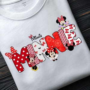 May include: A white t-shirt featuring the word MINNIE in large, stylized letters. The letters are filled with red and white polka dot patterns, floral motifs, and red geometric shapes. Minnie Mouse characters are integrated into the design.