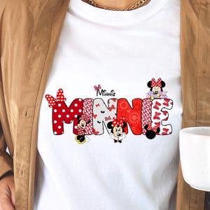 May include: A white t-shirt featuring a large graphic of the name MINNIE in bold, block letters. Each letter is filled with various red, white, and black patterns, including polka dots and bows, with small illustrations of Minnie Mouse integrated.