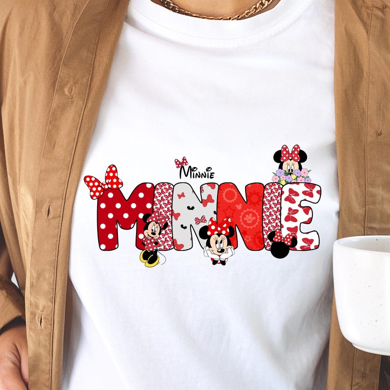 May include: A white t-shirt featuring a large graphic of the name MINNIE in bold, block letters. Each letter is filled with various red, white, and black patterns, including polka dots and bows, with small illustrations of Minnie Mouse integrated.