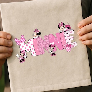 Puede incluir: Un art&iacute;culo de tela beige con el nombre "MINNIE" en letras rosas. Las letras est&aacute;n decoradas con lunares, lazos y personajes de Minnie Mouse. El art&iacute;culo est&aacute; sostenido por dos manos.
