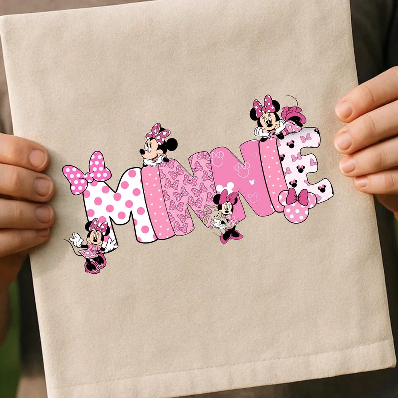 Puede incluir: Un art&iacute;culo de tela beige con el nombre "MINNIE" en letras rosas. Las letras est&aacute;n decoradas con lunares, lazos y personajes de Minnie Mouse. El art&iacute;culo est&aacute; sostenido por dos manos.