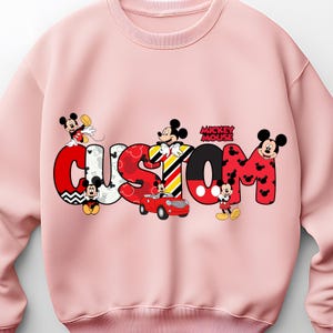 May include: A light pink sweatshirt featuring the word "CUSTOM" in a colorful, cartoonish font with Mickey Mouse characters incorporated into the design. The word "Mickey Mouse" is also printed on the sweatshirt.
