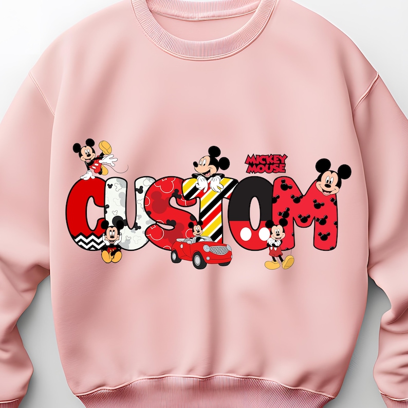 May include: A light pink sweatshirt featuring the word "CUSTOM" in a colorful, cartoonish font with Mickey Mouse characters incorporated into the design. The word "Mickey Mouse" is also printed on the sweatshirt.