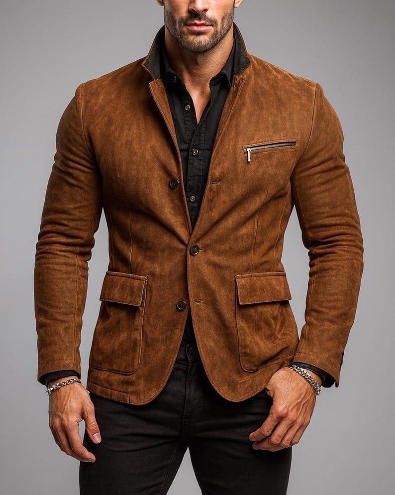 May include: A brown suede blazer with a black collar, a zippered chest pocket, and two front pockets. The blazer is buttoned and worn over a black shirt. The sleeves are long.