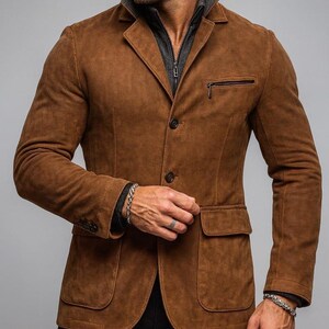 May include: A brown suede blazer with a notched lapel, two front pockets, and a zippered chest pocket. The blazer has a two-button closure and is worn over a gray zip-up jacket. The sleeves are long and the blazer is paired with black pants.