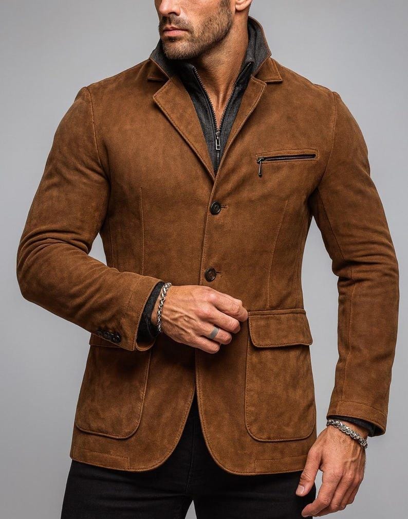 May include: A brown suede blazer with a notched lapel, two front pockets, and a zippered chest pocket. The blazer has a two-button closure and is worn over a gray zip-up jacket. The sleeves are long and the blazer is paired with black pants.
