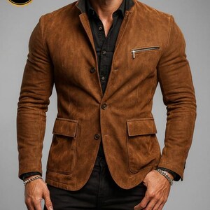 May include: A brown suede blazer with a black collar and a zippered chest pocket. The blazer has two front pockets and button closures. The sleeves are long and the fit is tailored. The blazer is worn over a black shirt.