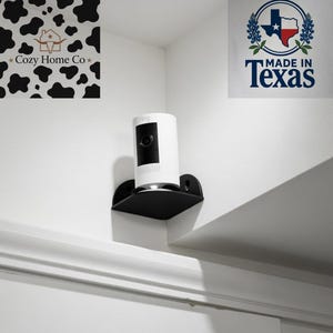 May include: A white security camera with a black lens is mounted on a black shelf. The shelf is attached to a white wall. The image also includes a "Made in Texas" logo and a "Cozy Home Co" logo.