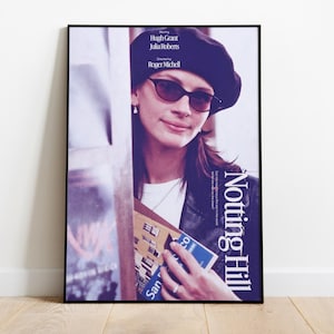 May include: A framed poster for the film "Notting Hill", featuring Julia Roberts in sunglasses and a beret. The poster is in a black frame and leans against a white wall. The poster includes the names of the actors and director.
