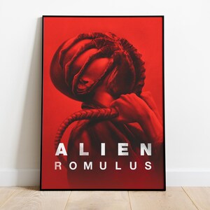 May include: Red poster in a black frame. The image is a stylized depiction of a figure with a large head and a rope-like element. White text reads "ALIEN ROMULUS". The poster is leaning against a white wall.
