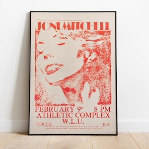May include: Framed poster featuring a red and cream-colored design. The poster displays the name "Joni Mitchell" at the top, with a stylized image of her face. Below, it lists the date, time, and location of a performance.