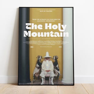 May include: A framed movie poster for The Holy Mountain featuring a central figure in white robes and a tall hat sitting between two goats. The background is yellow with black and white columns. Text includes the title and director Alejandro Jodorowsky.