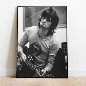 Peut inclure: Tirage encadré en noir et blanc d'un musicien jouant de la guitare. Le musicien porte un t-shirt à manches longues avec un logo de langue rouge. L'image est affichée sur un mur blanc au-dessus d'un sol en bois.