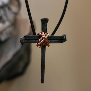 May include: A handcrafted cross pendant made from dark metal nails, bound with copper wire. The cross hangs from a dark leather cord. The background is a blurred neutral tone.