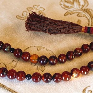 May include: A string of prayer beads with dark red, brown, and orange beads. The beads are strung together and have a dark brown tassel at the end. The beads are on a cream-coloured surface.