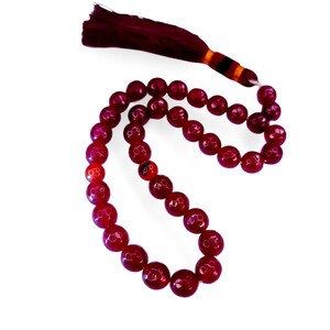 Puede incluir: Un collar o rosario de cuentas de piedras preciosas pulidas de color rojo oscuro. Las cuentas están ensartadas, con una borla a juego en la parte superior. Las cuentas tienen un acabado brillante y están colocadas sobre un fondo blanco.