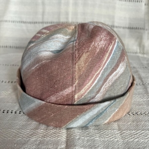 Puede incluir: Un sombrero estampado con un borde enrollado, que presenta una mezcla de colores. La tela tiene un diseño veteado en tonos rosa, marrón, azul y crema. El borde del sombrero es marrón liso y apagado.