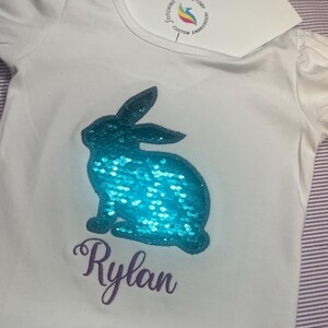 May include: White short-sleeved shirt with a turquoise sequined bunny design and the name "Rylan" in purple script. The shirt has a ruffled hem and puff sleeves. A card with a logo is placed on the shirt.
