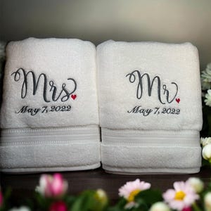 May include: Two white bath towels embroidered with "Mrs" and "Mr" in dark gray script, each with a small red heart. The date "May 7, 2022" is embroidered below the names. The towels are decorated with a horizontal stripe design.