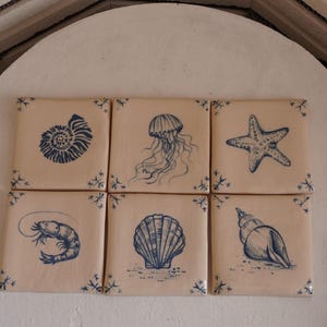 May include: Six square ceramic tiles with blue sea creature illustrations. The tiles feature a nautilus, jellyfish, starfish, shrimp, seashell, and conch shell. Each tile has a light beige background with blue corner accents.