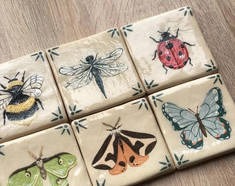 May include: A set of six square ceramic tiles featuring hand-painted insect illustrations. The collection includes a bumblebee, a dragonfly, a ladybug, a green moth, a brown and orange moth, and a blue butterfly, all on a cream background.