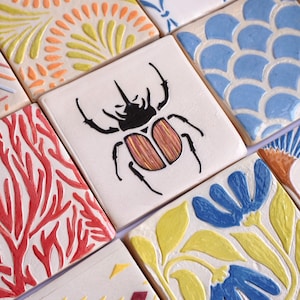 Peut inclure: Une collection de carreaux de céramique carrés présentant divers motifs en relief, notamment un scarabée noir, des branches de corail rouge, des fleurs bleues à feuilles vertes, des motifs en éventail orange et jaune, des écailles bleues et des formes géométriques rouges et jaunes.